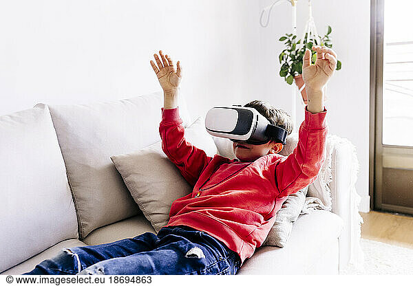Boy with arms raised wearing virtual reality headset lying on sofa at home