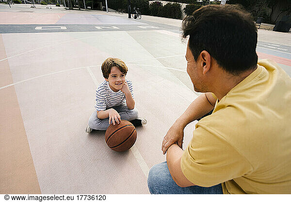 Boy with basketball sitting in front of father at sports court