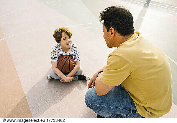 Boy with basketball sitting in front of father at sports court on sunny day