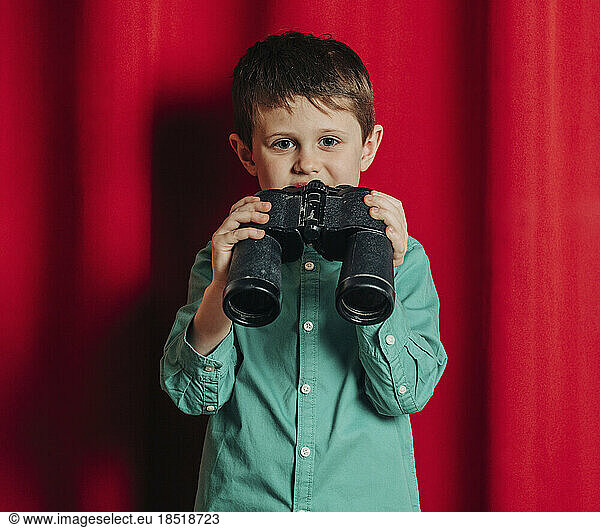Boy with binoculars standing in front of red curtain