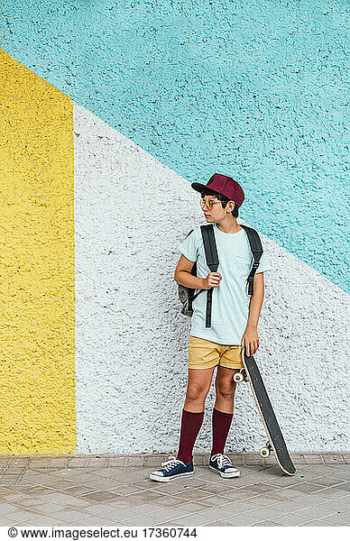 Boy with cap and skateboard standing on footpath