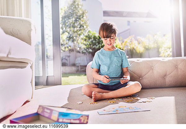 Boy with digital tablet assembling jigsaw puzzle on sunny living room floor