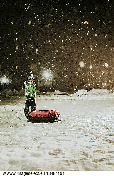Boy with inflatable sled standing in snow at night