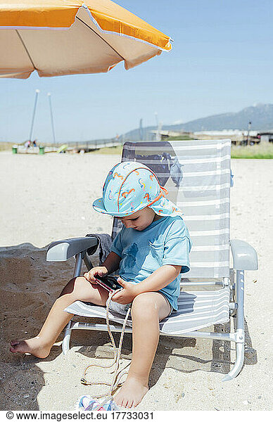 Boy with smart phone sitting on deck chair at beach