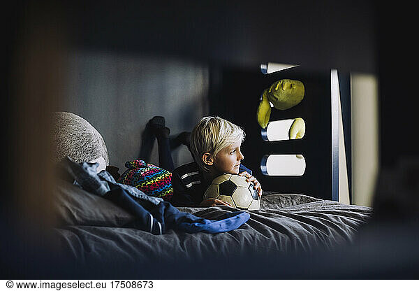 Boy with soccer ball looking away while lying on bed