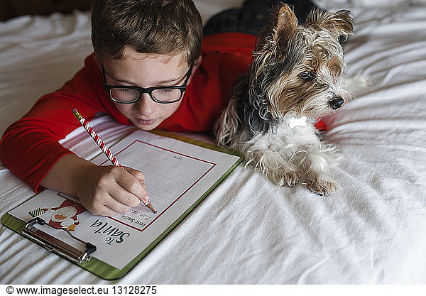 Boy writing on clipboard while lying on bed with Yorkshire Terrier