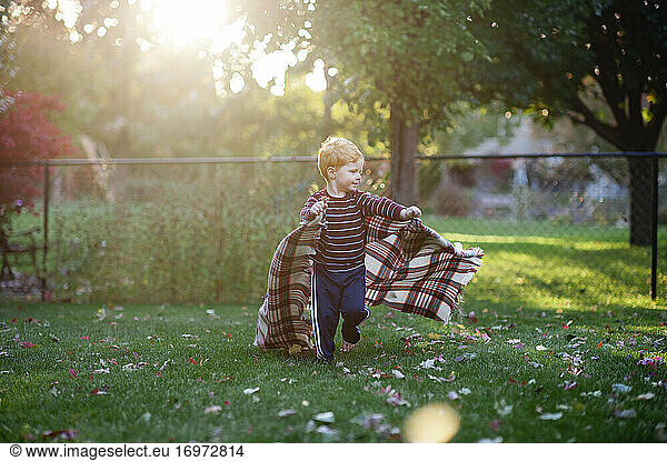 Boy 3-4 years old running backyard with blanket in pretty light
