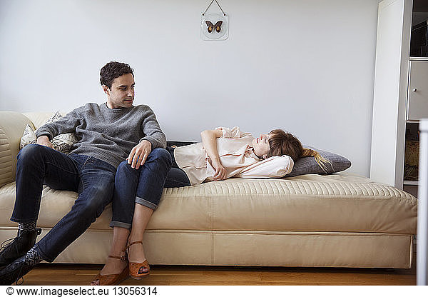 Boyfriend sitting by woman lying on couch against wall at home