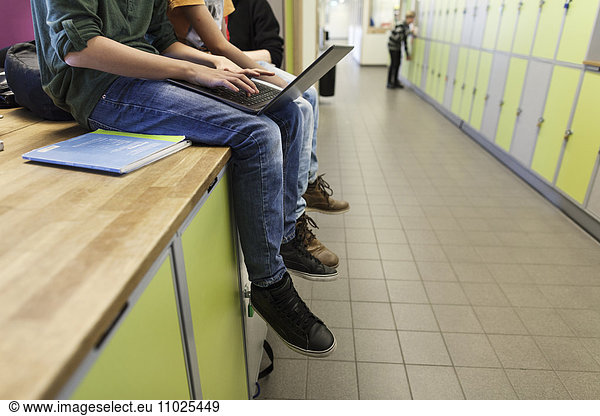 Boys (12-13) using laptop and sitting on lockers in school corridor