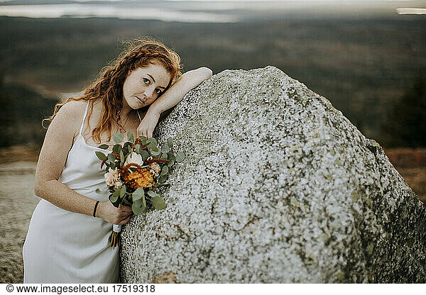 Bride with red hair and wedding flowers leans against boulder