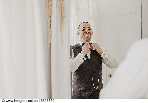 Bridegroom adjusting bow while looking in mirror at home