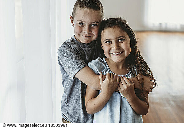 Brother and sister embracing in natural-light studio smiling at camera