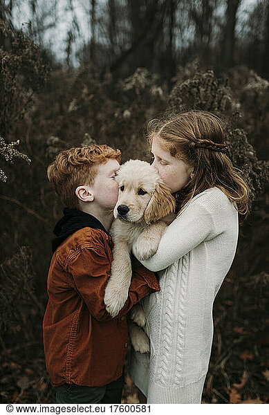 Brother and sister kissing golden retriever puppy in forest