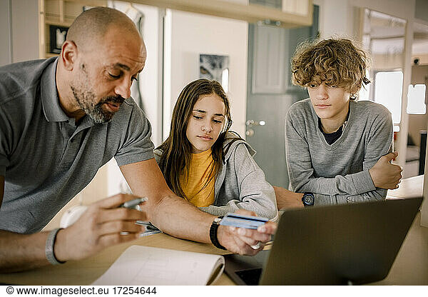 Brother and sister looking at father explaining finance with credit card at kitchen island