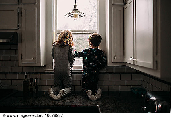Brother and sister looking outside kitchen window on cloudy day