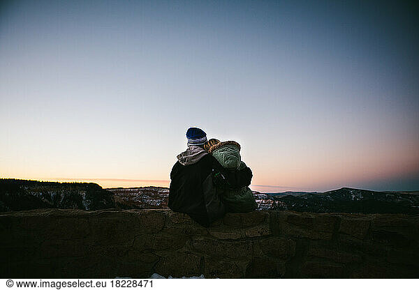 Brother and sister overlook snowy canyon in winter coats at sunset