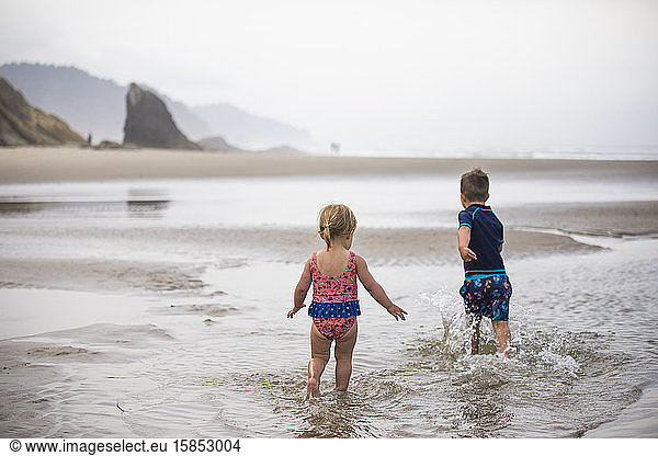 Brother and sister run through shallow water at the beach.