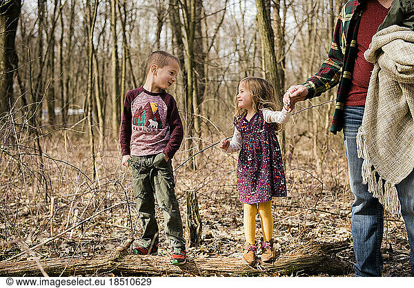Brother and sister smile at each other in woods holding dad's hand