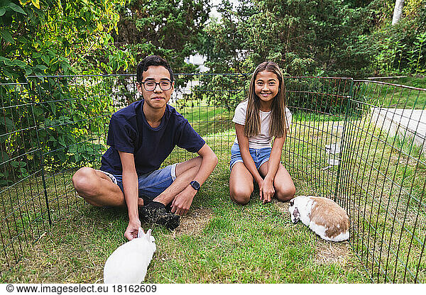 Brother and sister spending leisure time with rabbits amidst fence in backyard