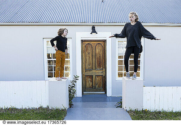 Brother and sister standing on a low wall outside a house laughing