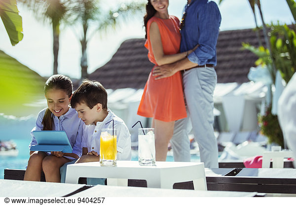 Brother and sister using digital tablet by resort swimming pool  parents embracing in background
