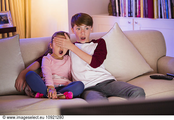 Brother covering surprised sister’s eyes watching TV in living room