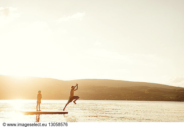 Brother diving into lake while girl standing on floating platform against sky during sunset