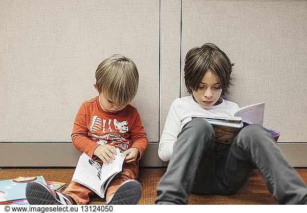 Brothers reading books while sitting on floor in library