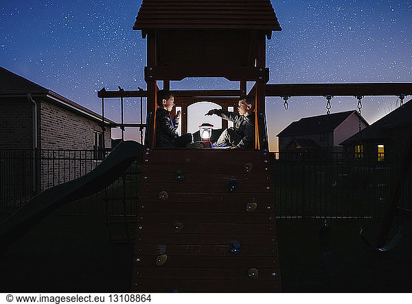 Brothers with illuminated lantern sitting on outdoor play equipment against star field at park