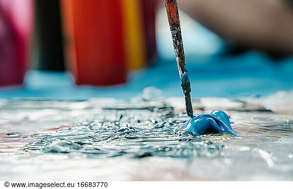 brush taking a blue color painting from a painting palette with some painting pots behind. Horizontal photo