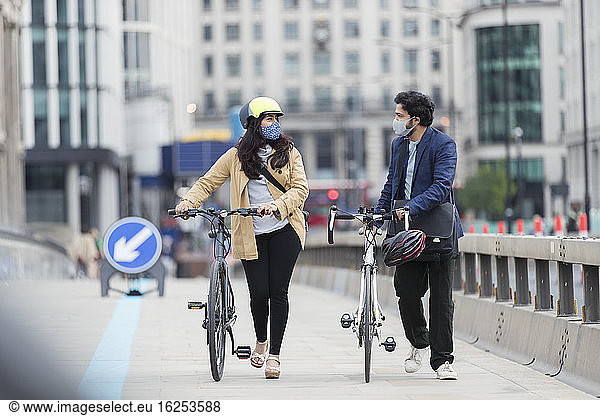 Business people in face masks walking bicycles on urban footbridge