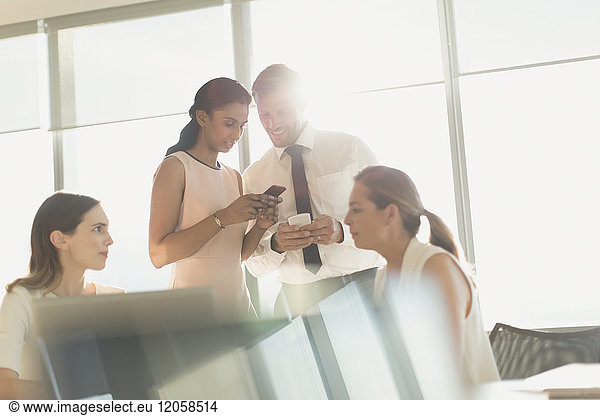 Business people texting with cell phones in sunny conference room meeting