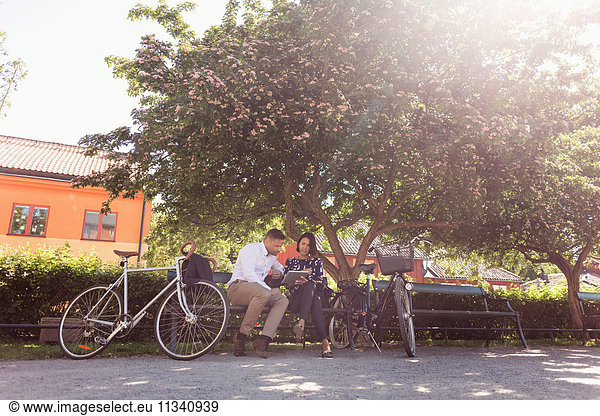 Business people using digital tablet while sitting on park bench by bicycles
