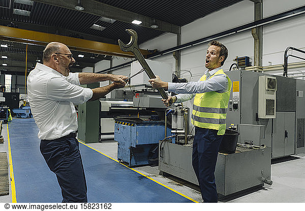 Businessman and man in reflective vest playfighting with large wrenches in a factory