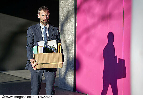 Businessman carrying box of personal belongings by wall on footpath