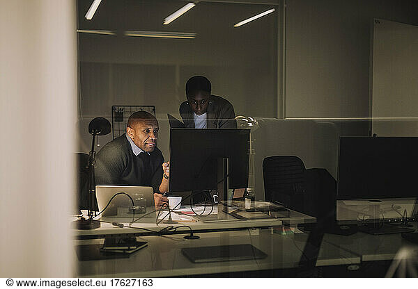 Businessman discussing with female colleague over computer seen through glass at work place