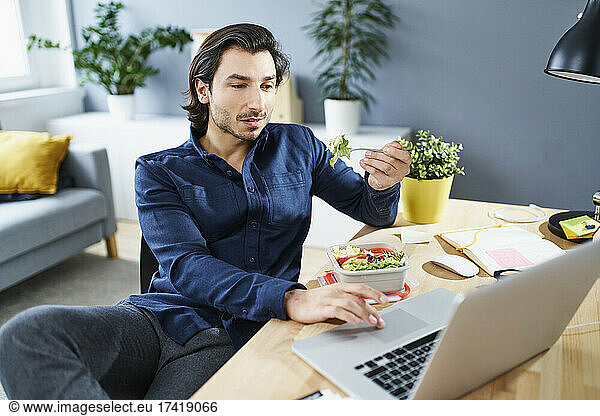 Businessman eating lunch while working on laptop at home