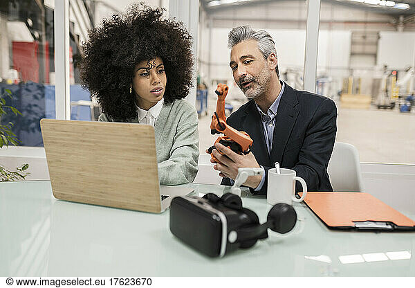 Businessman explaining robotic arm to colleague sitting at desk in factory