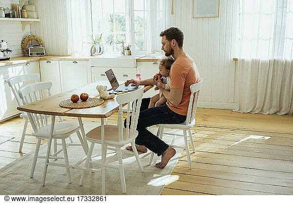 Businessman feeding daughter while working on laptop at home