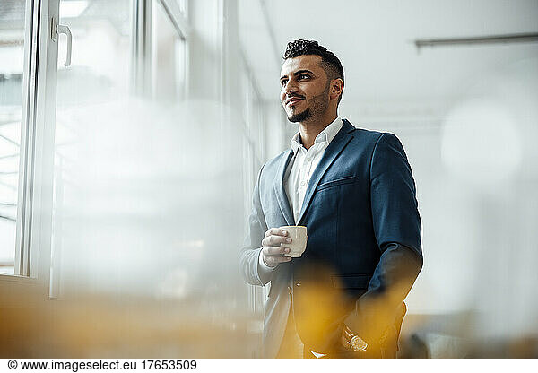 Businessman holding coffee cup looking through window standing in office