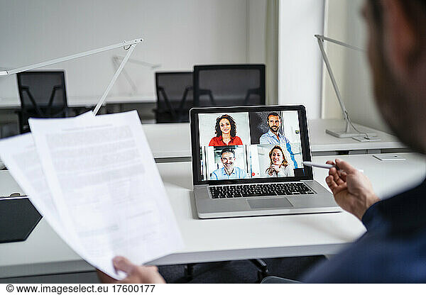 Businessman holding documents in discussion with colleagues on video call through laptop at office