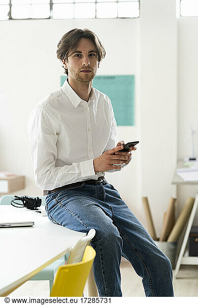 Businessman holding mobile phone while sitting on desk in creative office