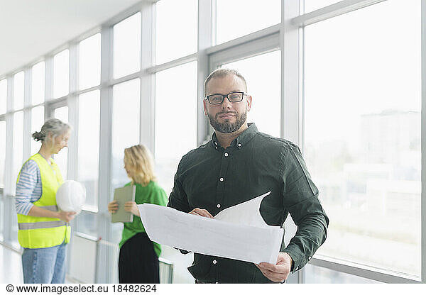 Businessman holding plan on office floor with colleagues in background