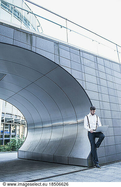 Businessman leaning against modern architecture and working on laptop  Munich  Bavaria  Germany