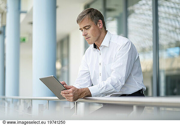 Businessman leaning on railing in office building using digital tablet