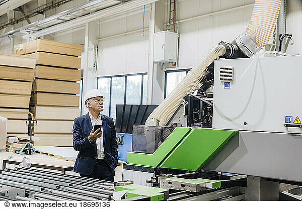 Businessman looking at modern machine holding smart phone in factory