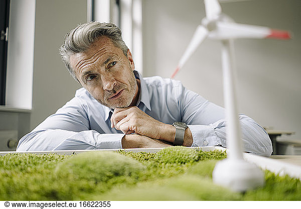 Businessman looking at wind turbine toy over moss frame while leaning on table at office