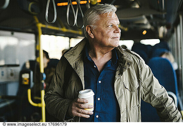 Businessman looking away holding disposable cup while standing in bus