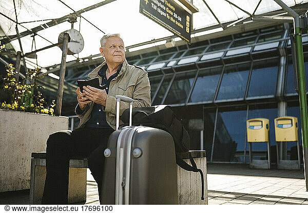 Businessman looking away holding smart phone while sitting on bench by luggage at railroad station