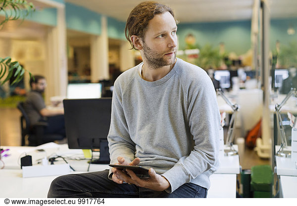 Businessman looking away while holding digital tablet on desk in creative office